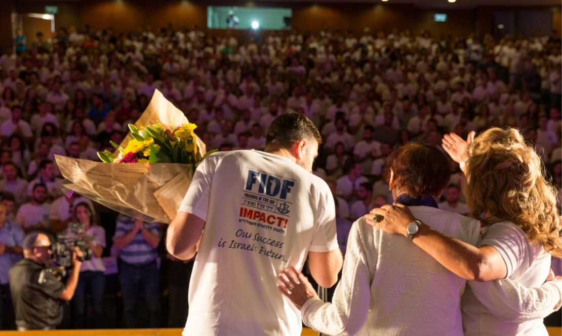 Man and two women on a stage, back to the camera, man wearing FIDF IMPACT! logo on his shirt and holding a bouqet of flowers, audience in the background